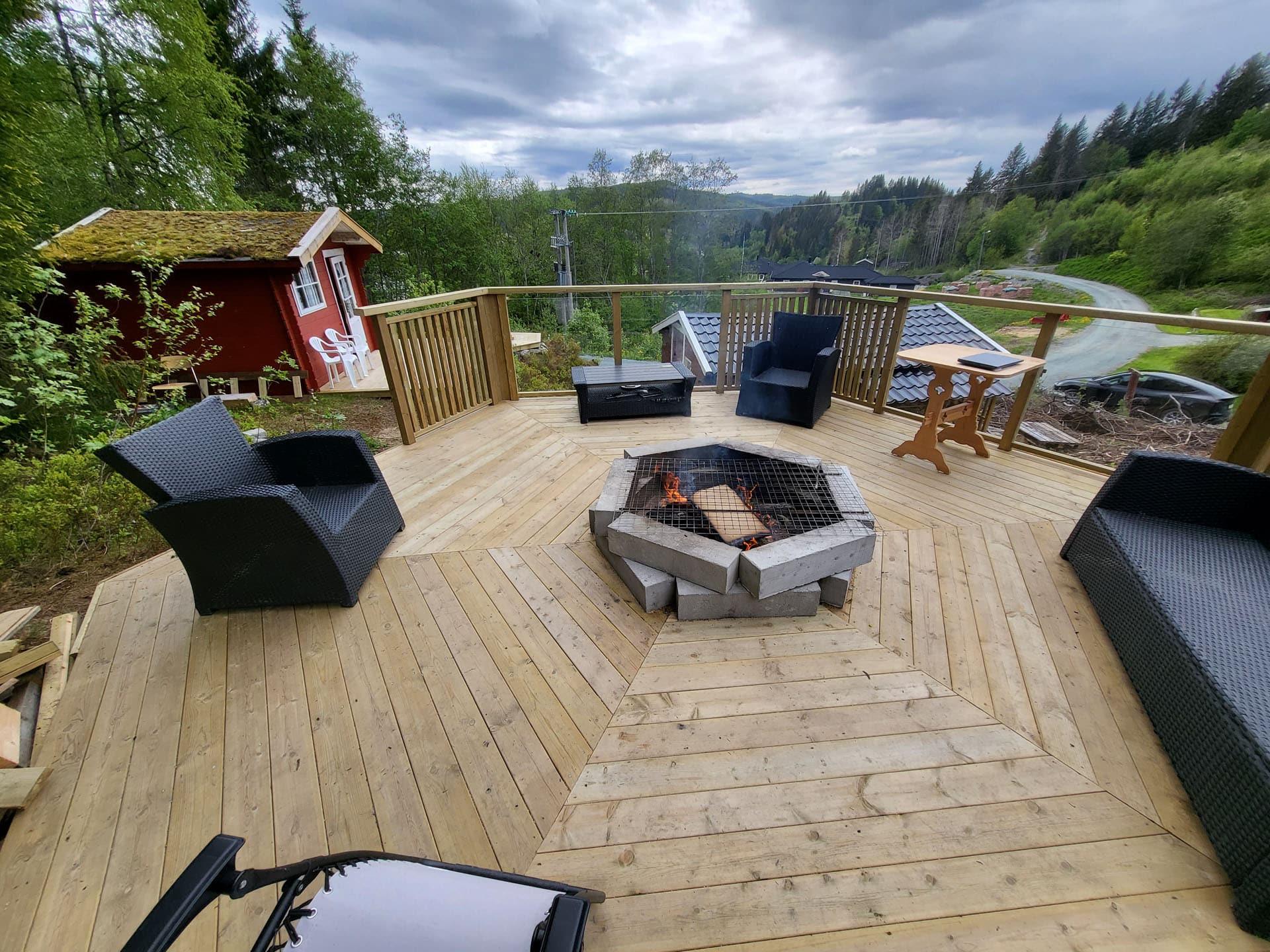 Stor terrasse i tre med bålpanne og utemøbler i skogkanten
